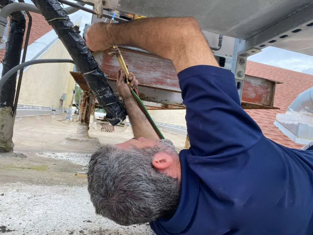 Technician lying under a rooftop AC unit, working on its lines with a torch